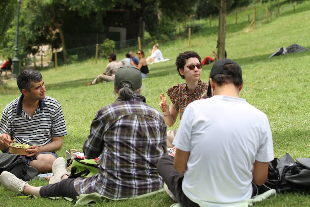 Membres du GEM TSA/TND de Paris réunis dans un parc pour un pique-nique, discutant et partageant un moment chaleureux en plein air, illustrant l’inclusion, la pair-aidance, l’autonomie et la vie sociale au sein du GEM.